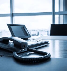 A close-up photo of a modern VoIP phone handset placed on a sleek office desk. The phone has a large touchscreen display and several buttons for functions like mute, hold, and speaker. Behind the phone, a blurred cityscape is visible through a window.