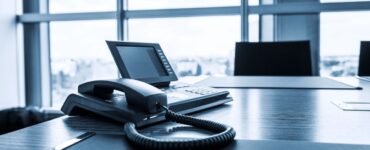 A close-up photo of a modern VoIP phone handset placed on a sleek office desk. The phone has a large touchscreen display and several buttons for functions like mute, hold, and speaker. Behind the phone, a blurred cityscape is visible through a window.