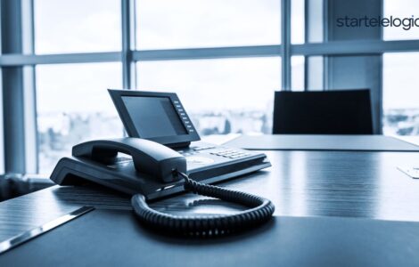 A close-up photo of a modern VoIP phone handset placed on a sleek office desk. The phone has a large touchscreen display and several buttons for functions like mute, hold, and speaker. Behind the phone, a blurred cityscape is visible through a window.